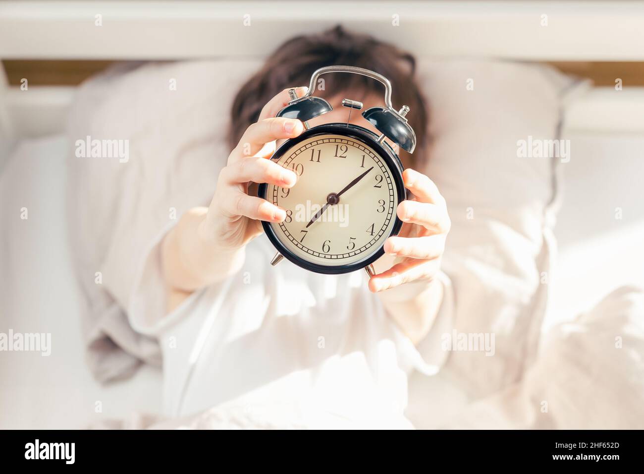 Unrecognizable kid lying in bed and holding a retro style alarm clock. Time to get up in the morning. Focus on the clock Stock Photo