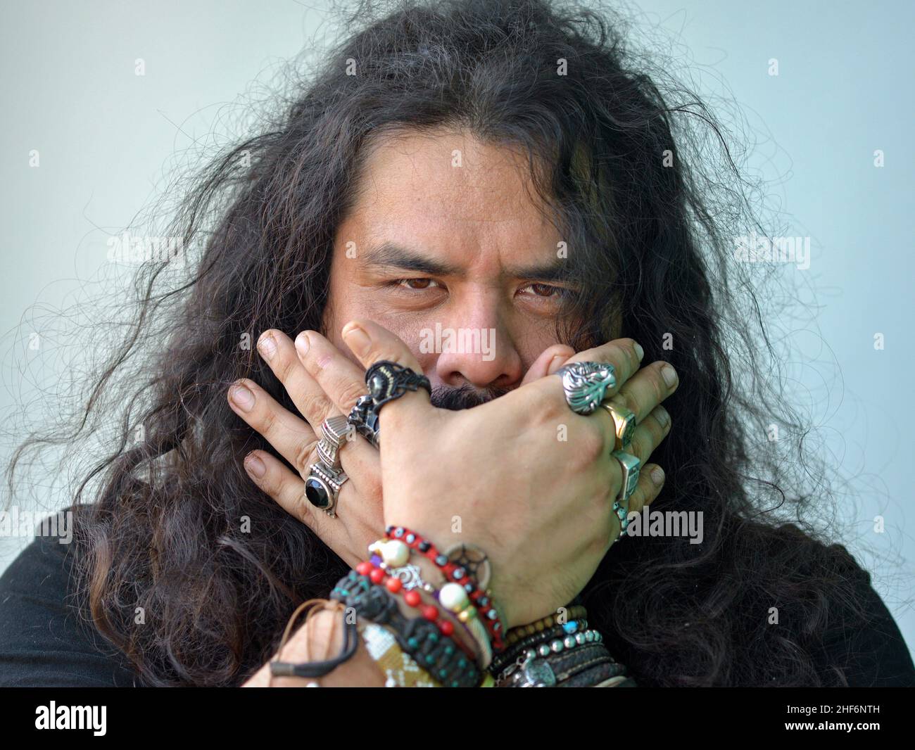 Young Caucasian man with long hair and beringed hands expresses speak-no-evil (iwazaru) of proverbial principle of Three Wise Monkeys (one of three). Stock Photo