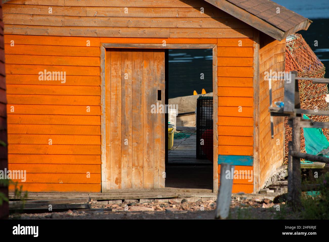 Orange wooden fishing shed for storing fishing gear and boats. The building has a worn wooden door, window, peaked roof, fishing rope, and gear. Stock Photo