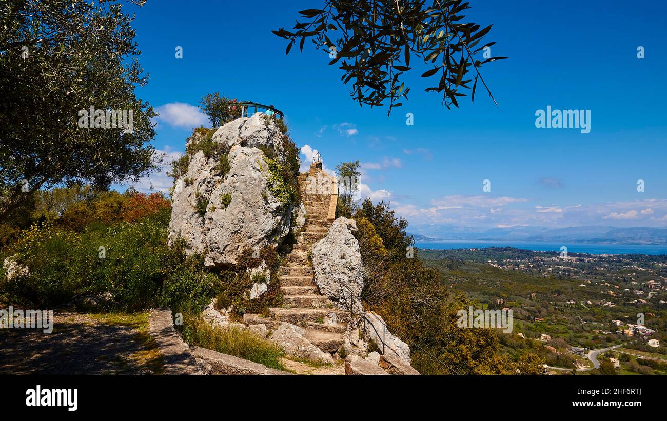Greece,  Greek Islands,  Ionian Islands,  Corfu,  central north of the island,  viewpoint "Emperor's Throne",  view of the west and east coast,  view platform,  blue sky,  white clouds,  mountains on the mainland,  view to the round,  on a rock built observation deck,  stone stairs framed by the branches of an olive tree,  blue sky,  some white clouds Stock Photo