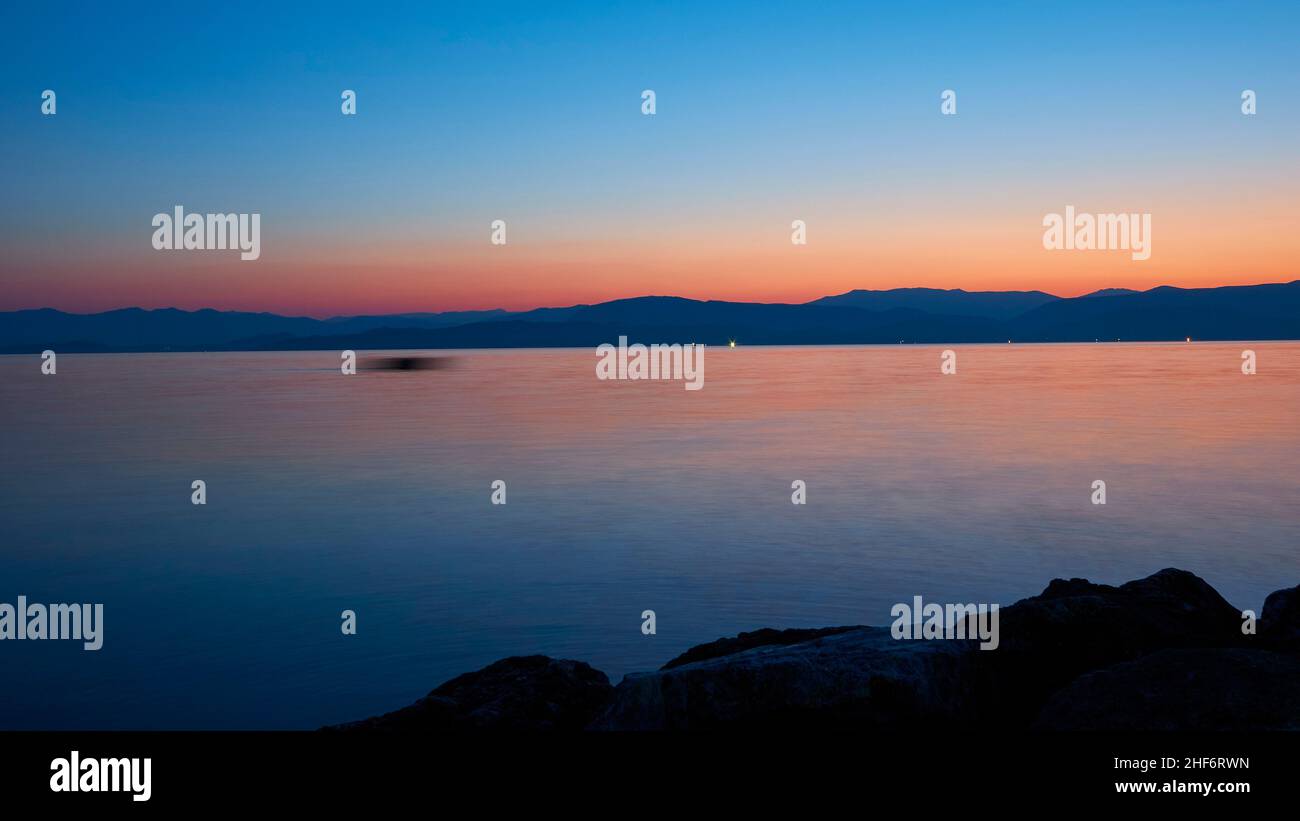 Greece,  Greek Islands,  Ionian Islands,  Corfu,  Corfu Town,  sunrise at the sea,  view over the sea to the hills of the mainland,  blue sky above,  below orange-red stripes,  calm sea,  blurred moving fishing boat in the middle distance Stock Photo