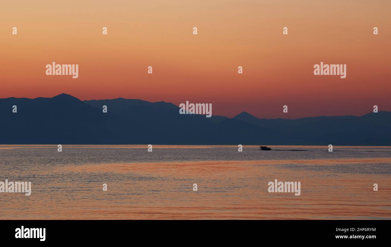 Greece,  Greek Islands,  Ionian Islands,  Corfu,  Corfu Town,  sunrise at the sea,  view over the sea to the hills on the mainland,  sky pink to red,  fishing boat moves from right to left in the middle distance Stock Photo