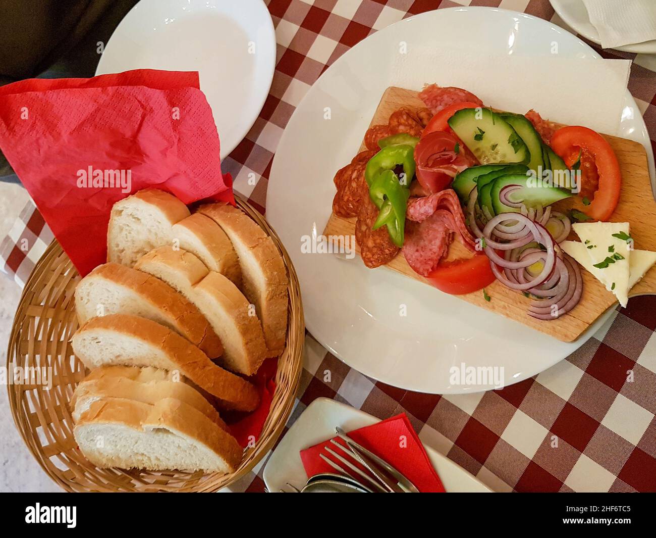 A selection of Italian meats with cheese, cucumber and chillies served on a wooden plater board with a bread basket to accompany the meal. Traditional Stock Photo