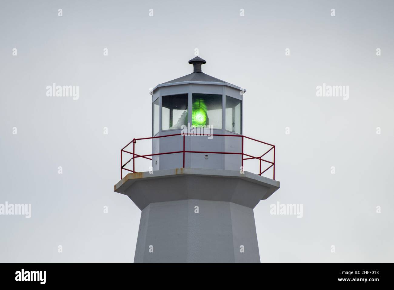 The watch room of a vintage eight-sided lighthouse. The lantern room has a large green bulb. The concrete building is white with a white railing. Stock Photo