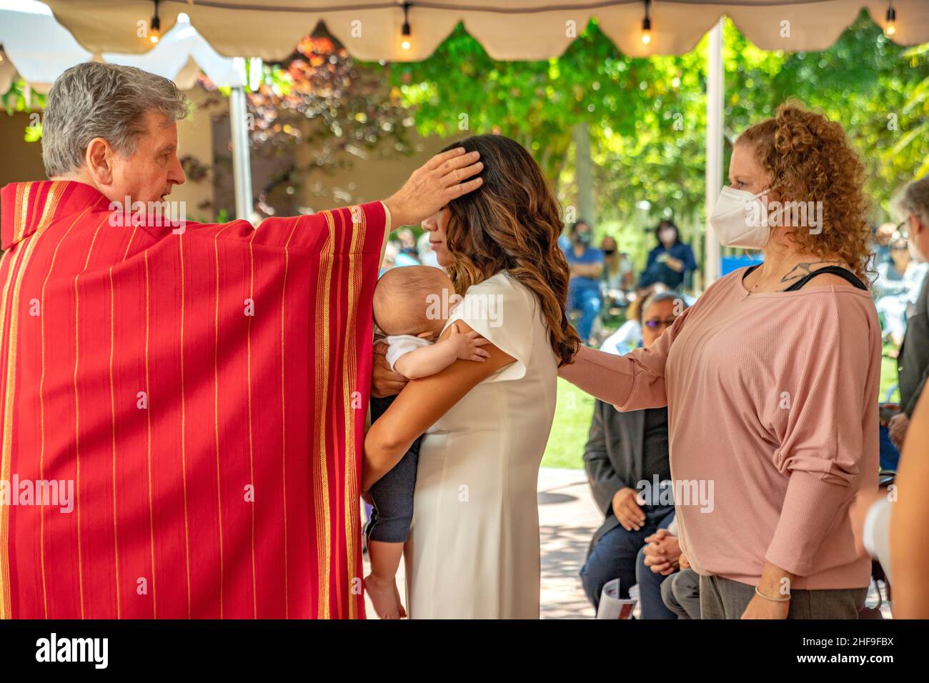 Holding her baby, a candidate for adult baptism is blessed by a monsignor at a Southern California Catholic church. Note sponsor at right. Stock Photo