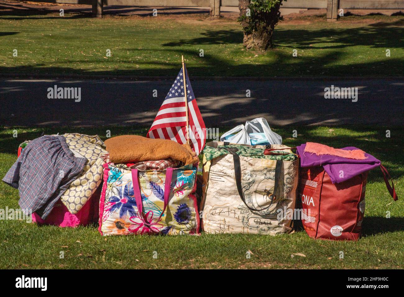 Tote bags holding possessions and clothing of a homeless man include a US flag in an Anaheim, CA, park where the owner will sleep outdoors. Stock Photo