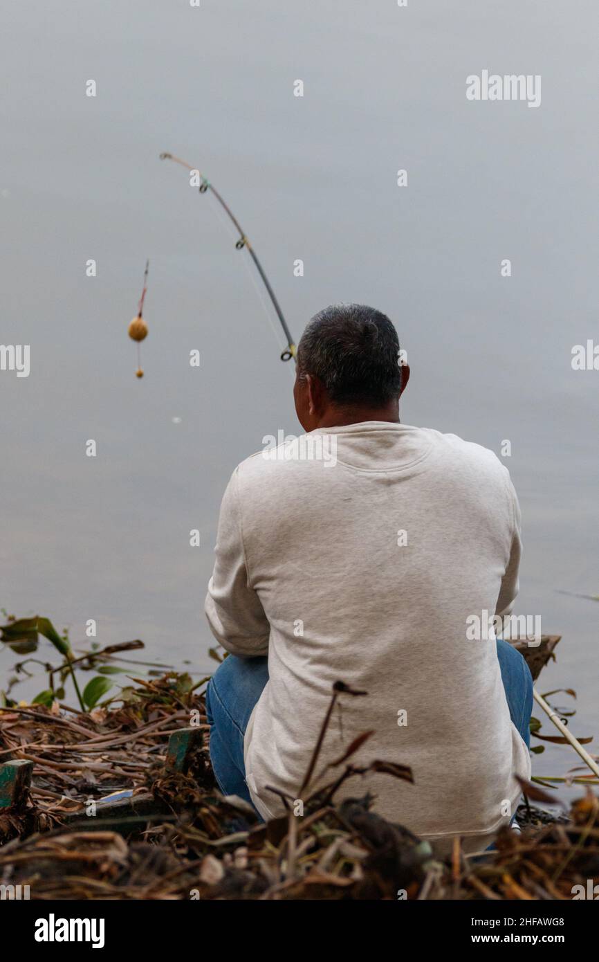 A lone fisherman sits on the banks of Phewa Lake with his pole in the water Stock Photo