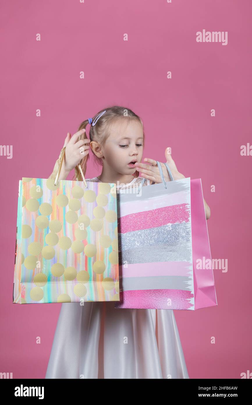 Shopping little caucasian girl in a party dress on pink background holding shopping bags Stock Photo
