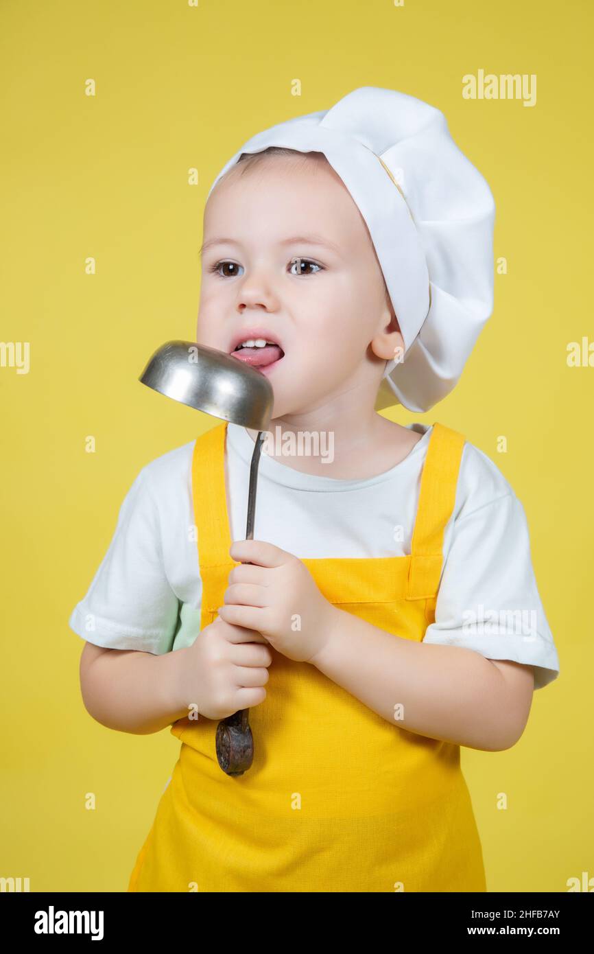 Little caucasian Boy playing chef, boy in apron and chef's hat licks the ladle on yellow background Stock Photo