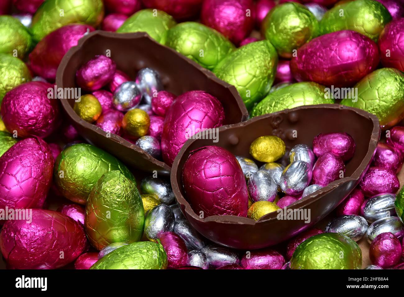 Pile of multi colored and different sizes of colourful foil wrapped chocolate easter eggs in pink, red, gold, yellow and lime green with two halves of Stock Photo