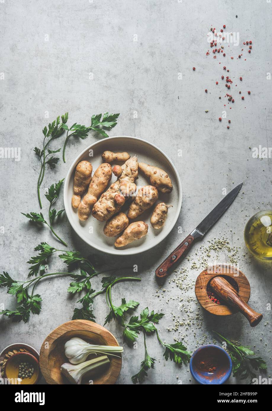 Jerusalem artichoke in bowl on grey concrete kitchen table with ingredients, parsley, mortar and pestle, kitchen knife, oil and garlic. Preparing vege Stock Photo