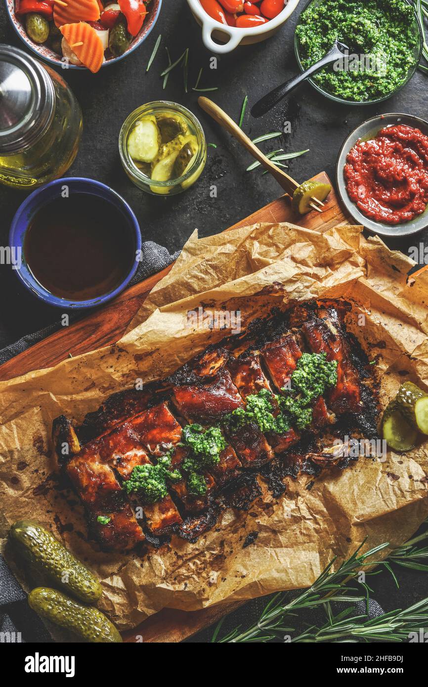 Roasted glazed bbq ribs with green pesto in baking paper at table with green sauce, salsa, oil and pickled cucumber. Eating grilled barbecue meat. Top Stock Photo