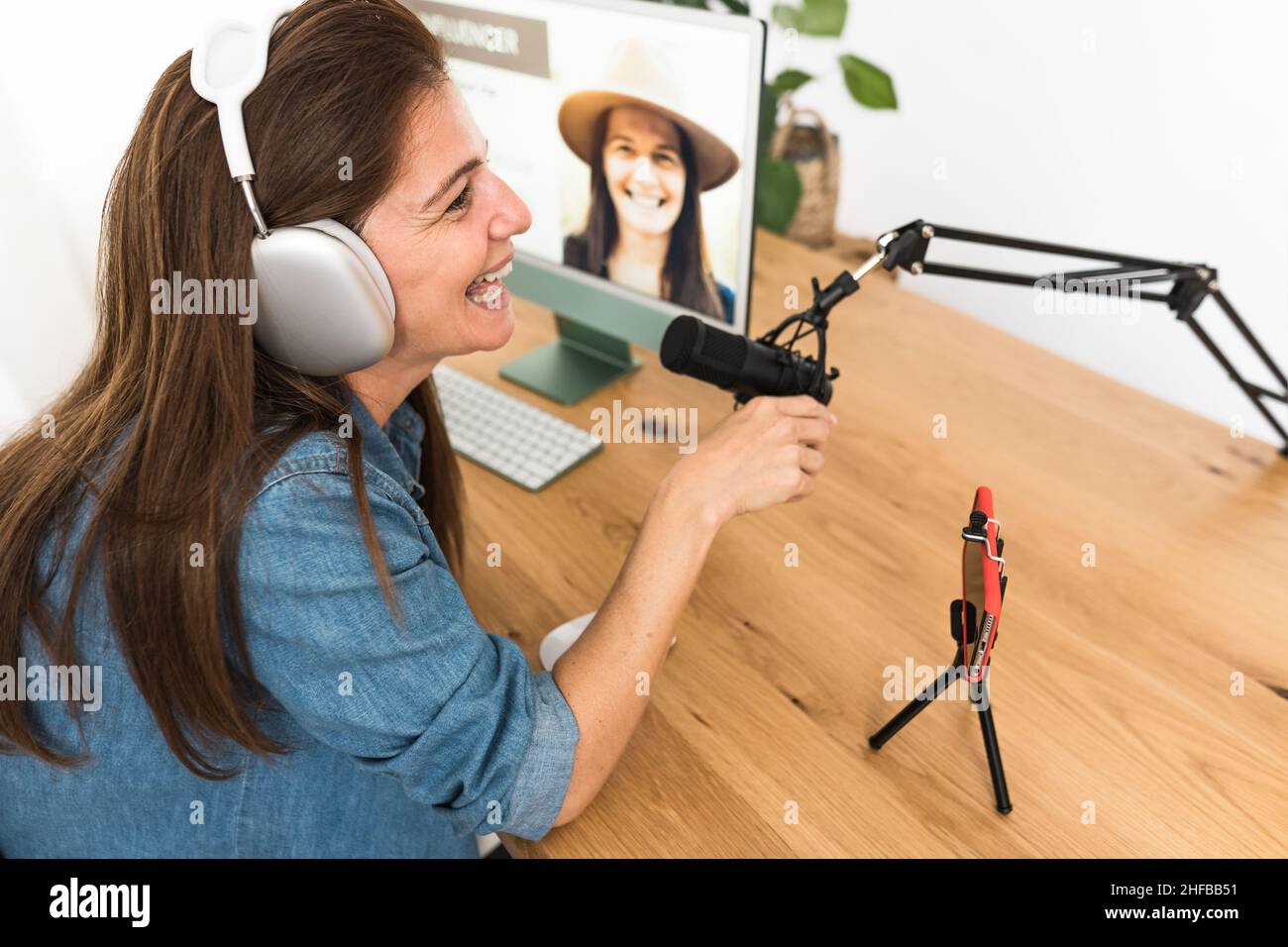 Mature woman recording a podcast using microphone and laptop while streaming on social network with mobile smartphone from her home studio Stock Photo