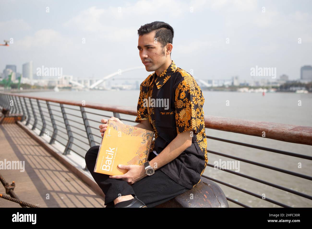 A young male model dressed in Indonesian Batik reads a book entitled Batik with the background of the Huangpu river and Lupu bridge at the Long Museum Stock Photo