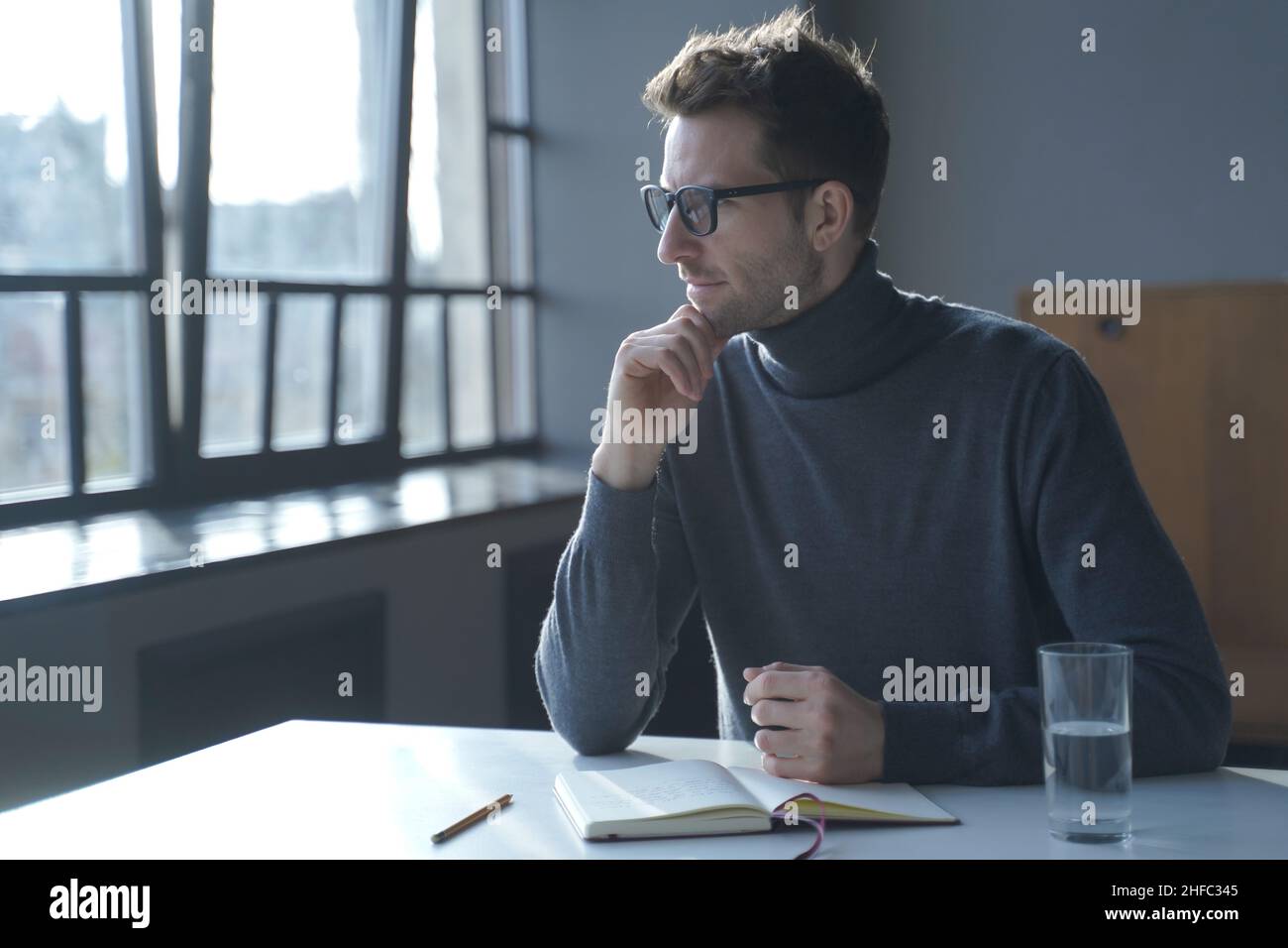 Young pensive thoughtful German man sitting at desk and looking out window Stock Photo