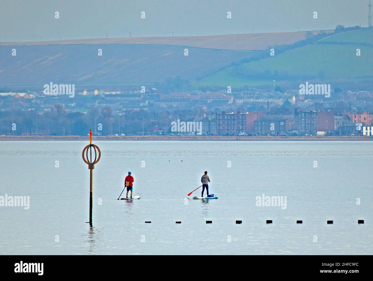 Portobello, Edinburgh, Scotland, UK. 15th January 2022.  Calm Firth of Forth for another cloudy afternoon, temperature of 7 degrees centigrade for those engaged in watersports. Pictured: Two males on paddleboards enjoy the calm conditions with East Lothian in the background. Credit: Archwhite/Alamy Live News. Stock Photo