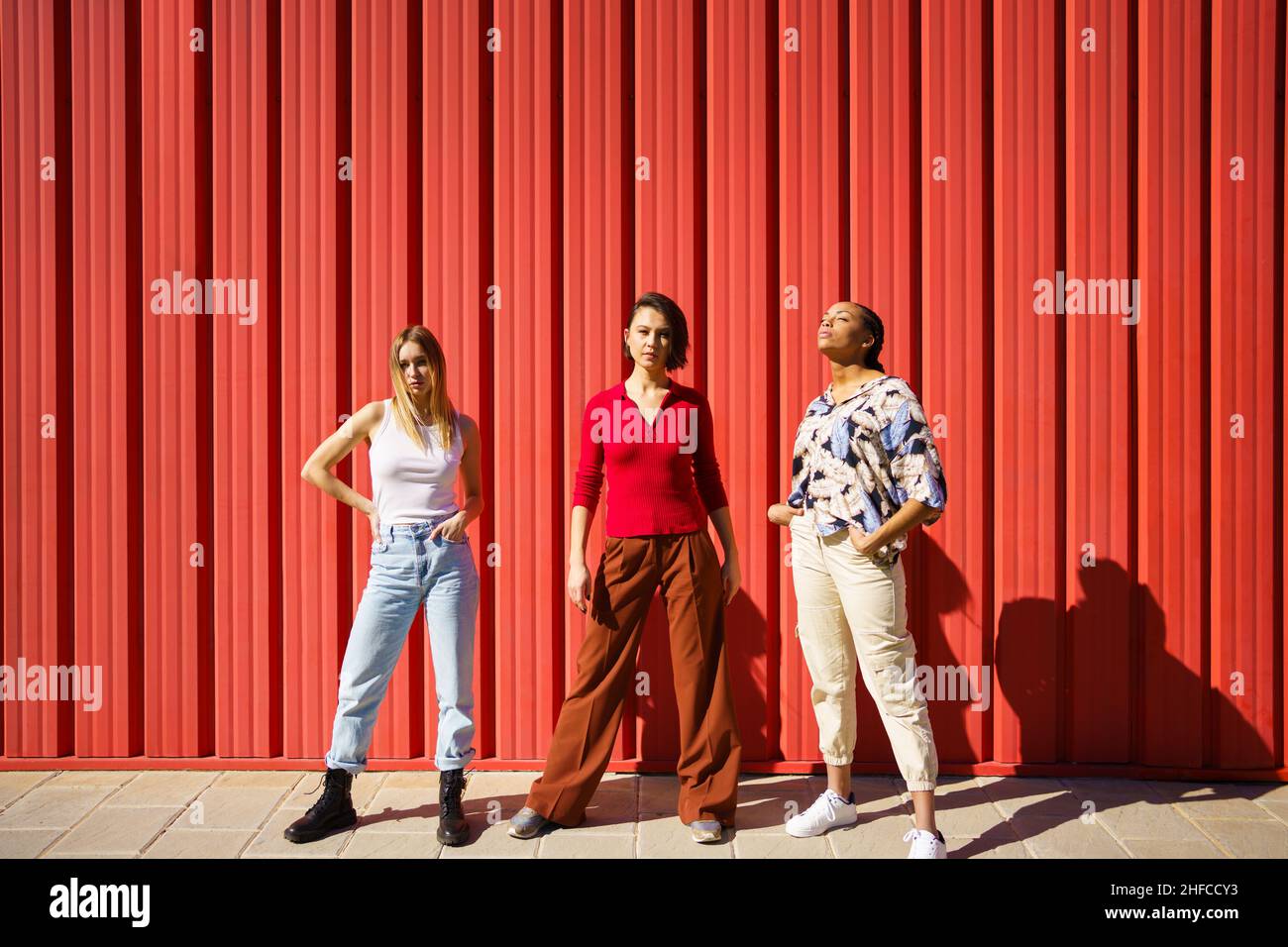 Stylish young diverse ladies standing against red fence on street Stock Photo