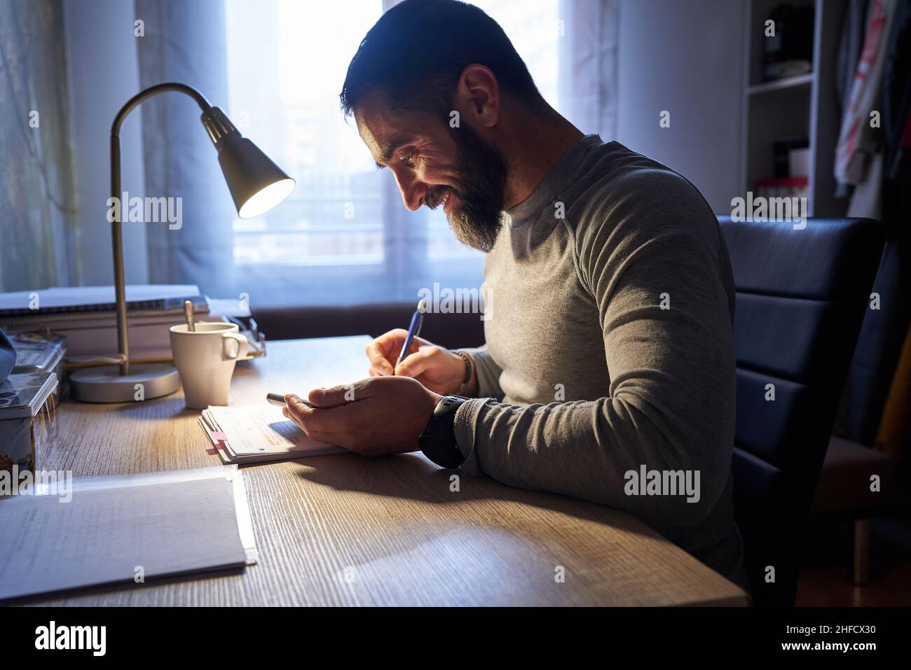 Young white man with a neat beard studying and looking at the phone at his desk Stock Photo