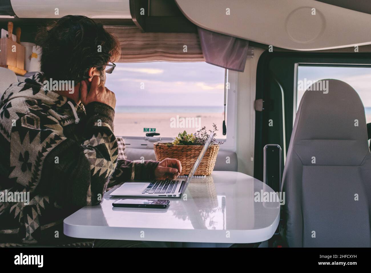 Man working inside camper van sitting at the table and looking outside the window the beautiful beach and ocean in background. Concept of freedom life Stock Photo