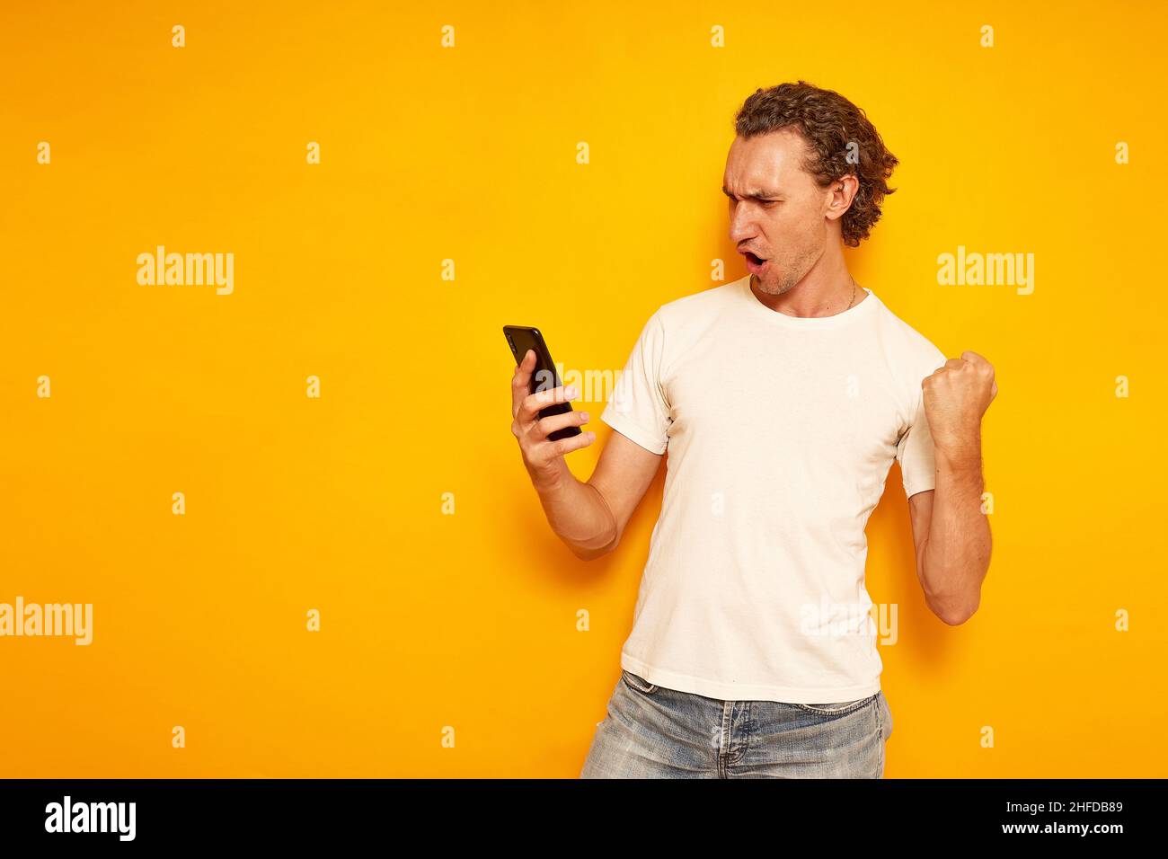 excited man checks his mobile phone celebrating good news with victorious gesture isolated on yellow background in casual clothes. space for text. Concept - communication, people, social networks. High quality photo Stock Photo