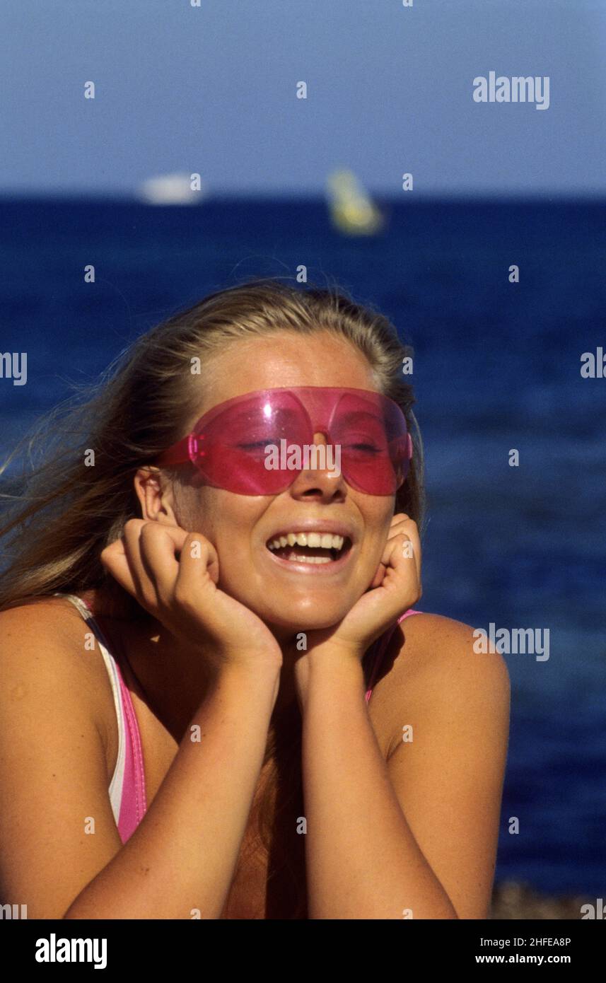 dynamic blond hair young woman beauty smiling front view camera pink sunglasses blue seawater background Stock Photo