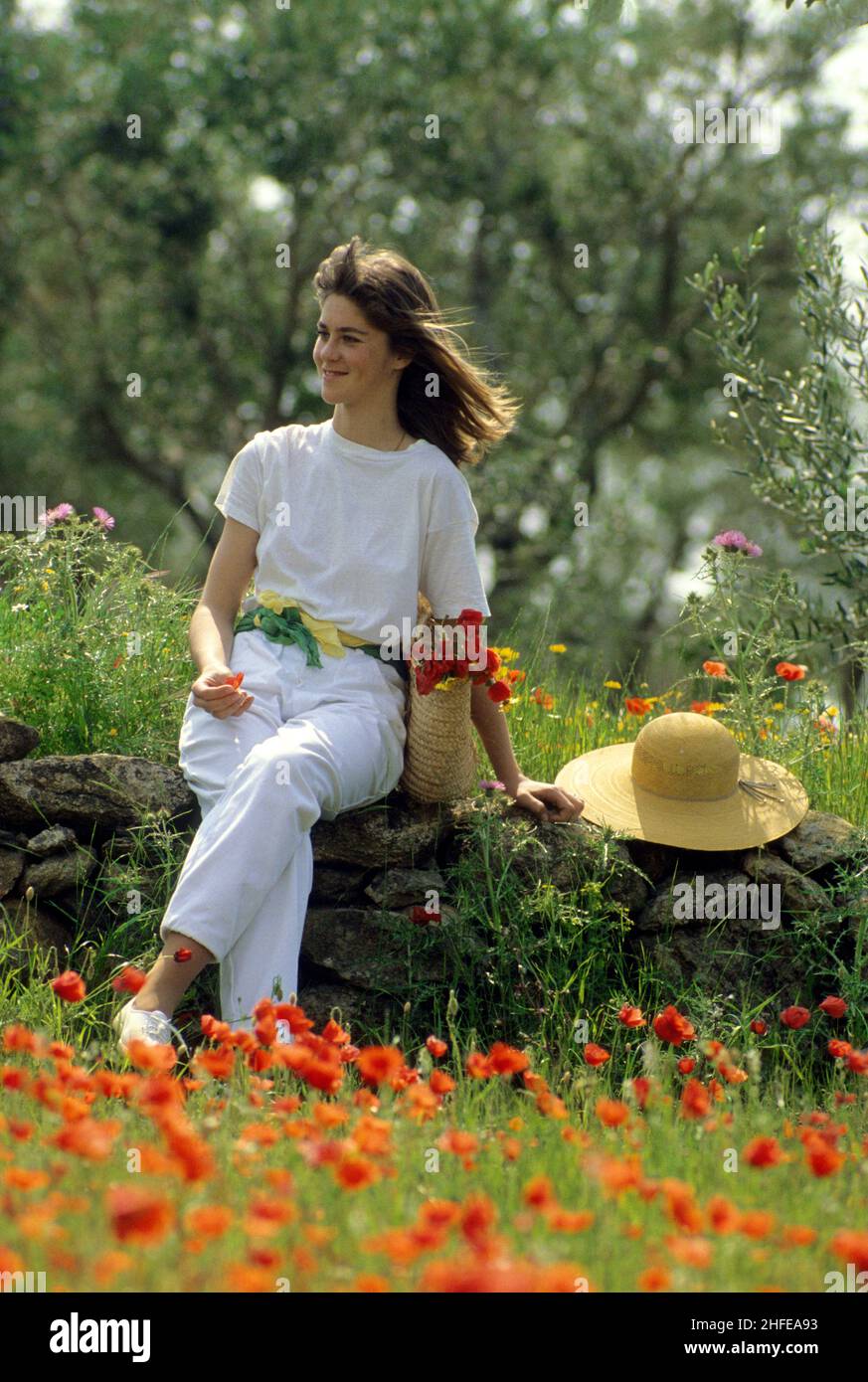 blond hair happy young girl sit on stone wall middle country field with poppies all white dressed spring time Stock Photo