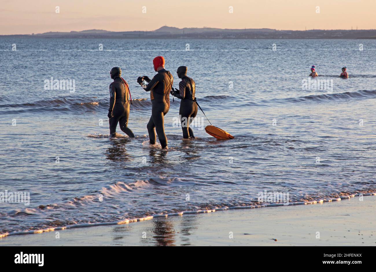 Portobello, Edinburgh, Scotland, UK. 16th January 2022. Finally sunrise sunshine with temperature of 7 degrees after a week of heavy cloud in the skies above the city. Pictured: Friends in wetsuits head into the chilly Firth of Forth for a swim. Credit: Scottishcreative/Alamy Live News. Stock Photo