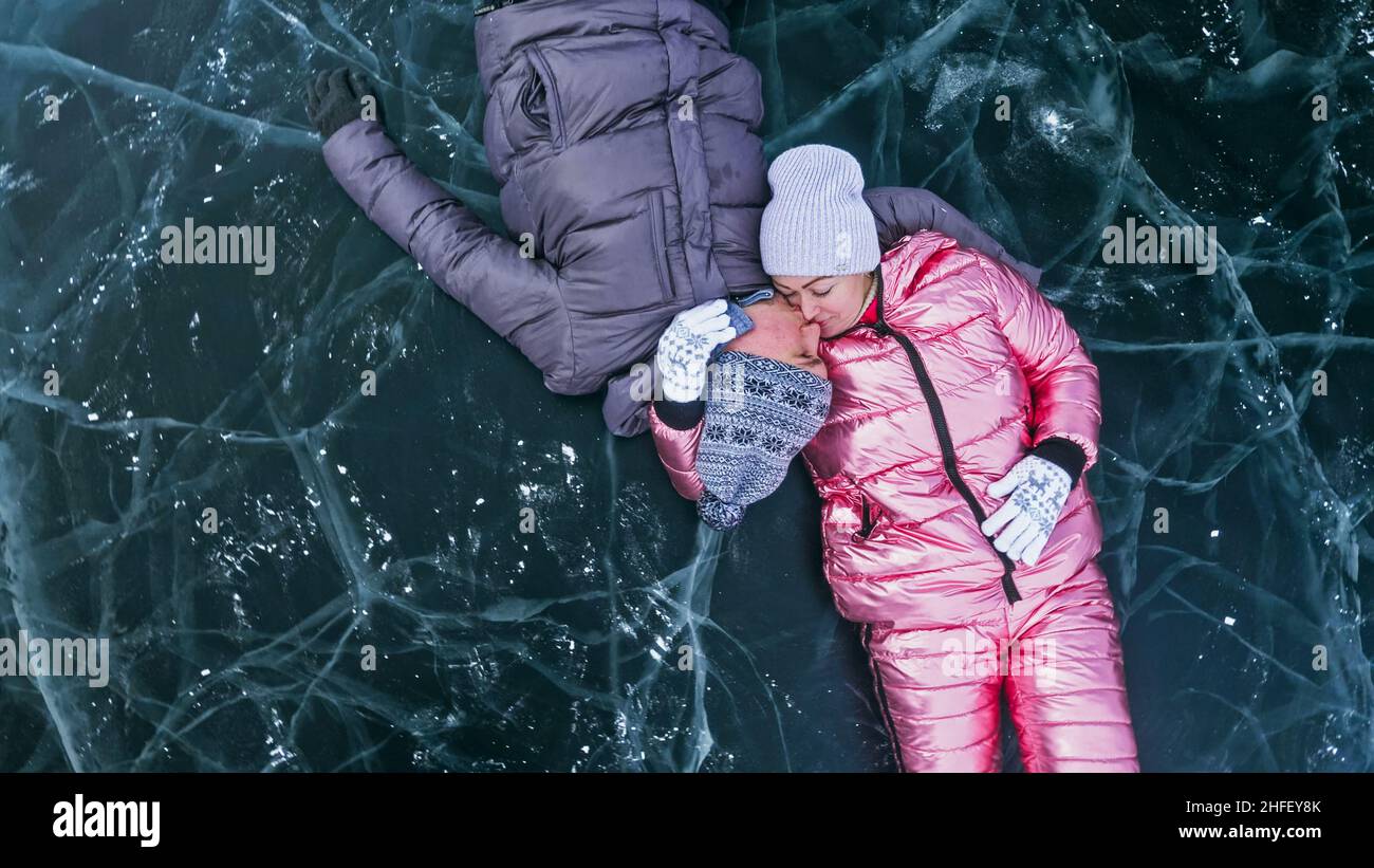 Couple has fun winter walk against background of ice of frozen lake. Lovers lie on clear ice with cracks have fun kiss and hug. View from above. Happy people on snow covered ice. Honeymoon love story. Stock Photo