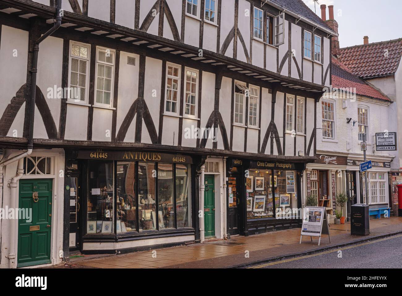 An ancient half-timbered building with a row of shops on the ground floor.  The pavement is wet from recent rain. Stock Photo