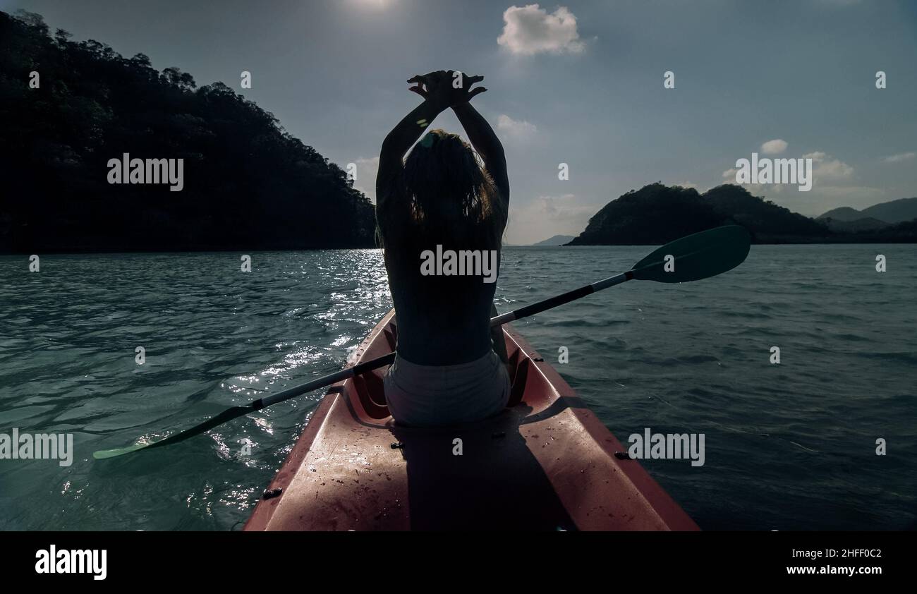 Silhouette of woman tourist raising hands while sailing in canoe along sea bay water to distant hilly island backside view. Stock Photo