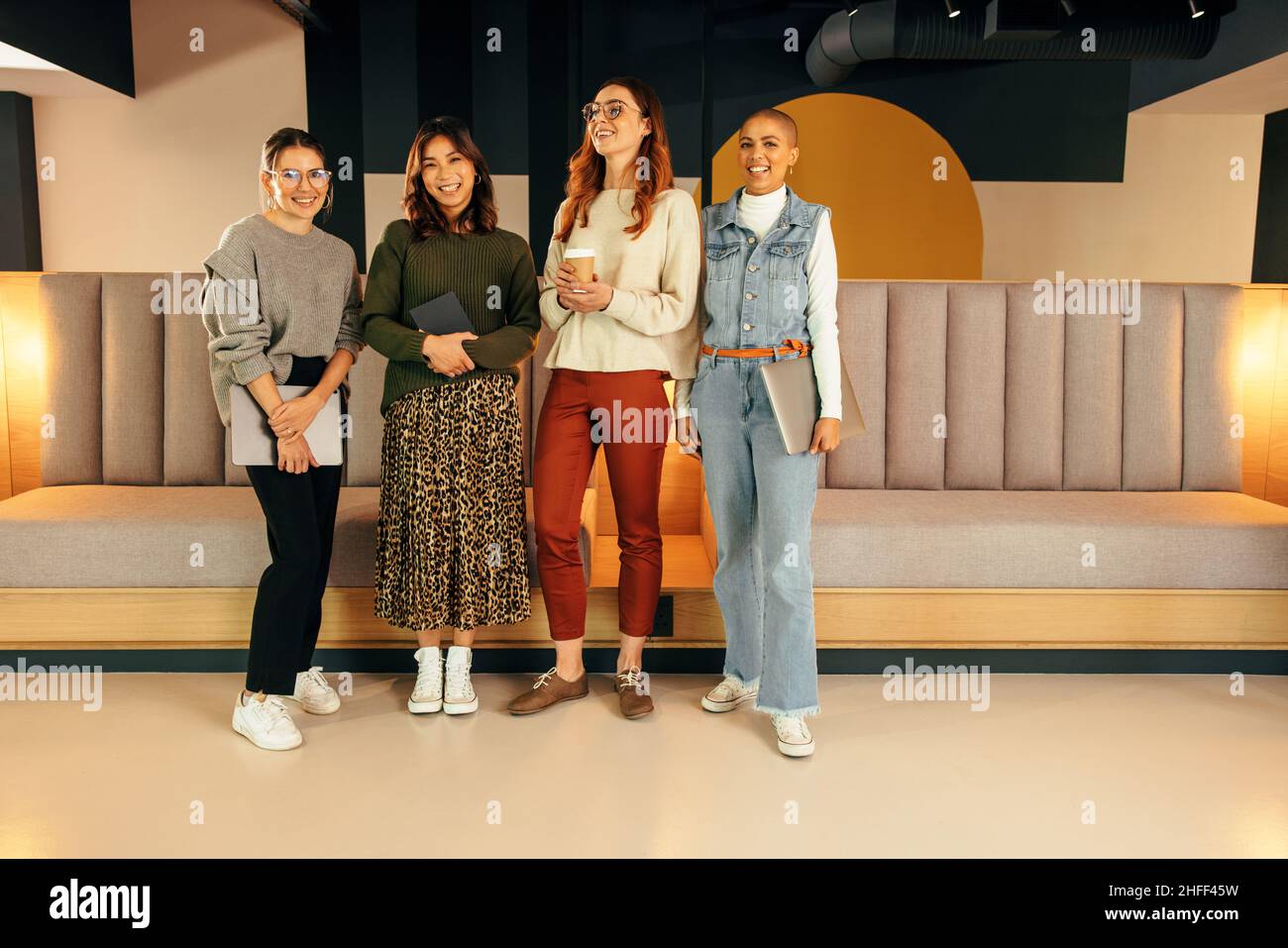 Four female entrepreneurs smiling happily while standing together in an office lobby. Group of multiethnic businesswomen working as a team in a modern Stock Photo
