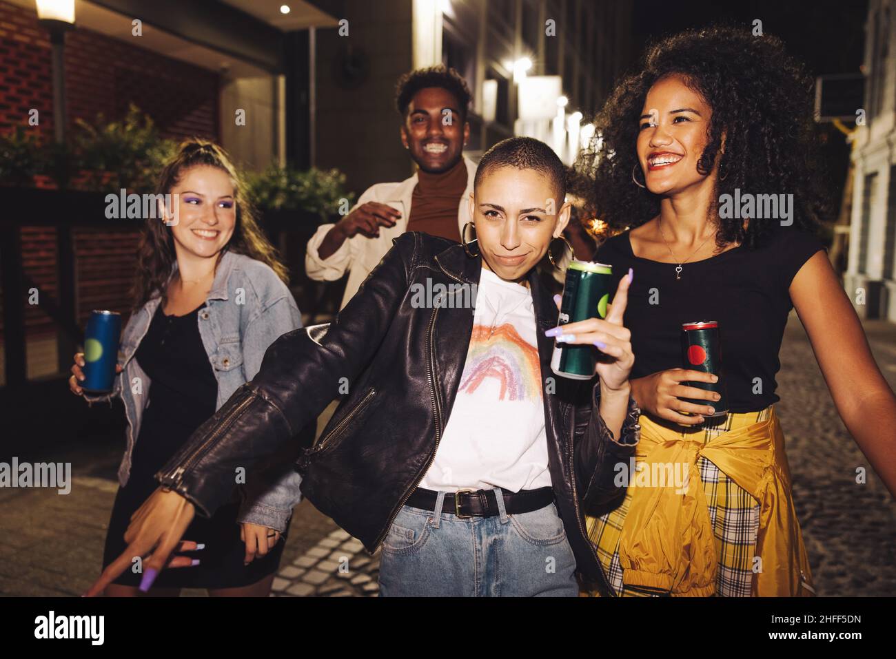 Group of happy friends dancing in the city at night. Four vibrant young people holding beer cans while walking together in the street. Friends having Stock Photo