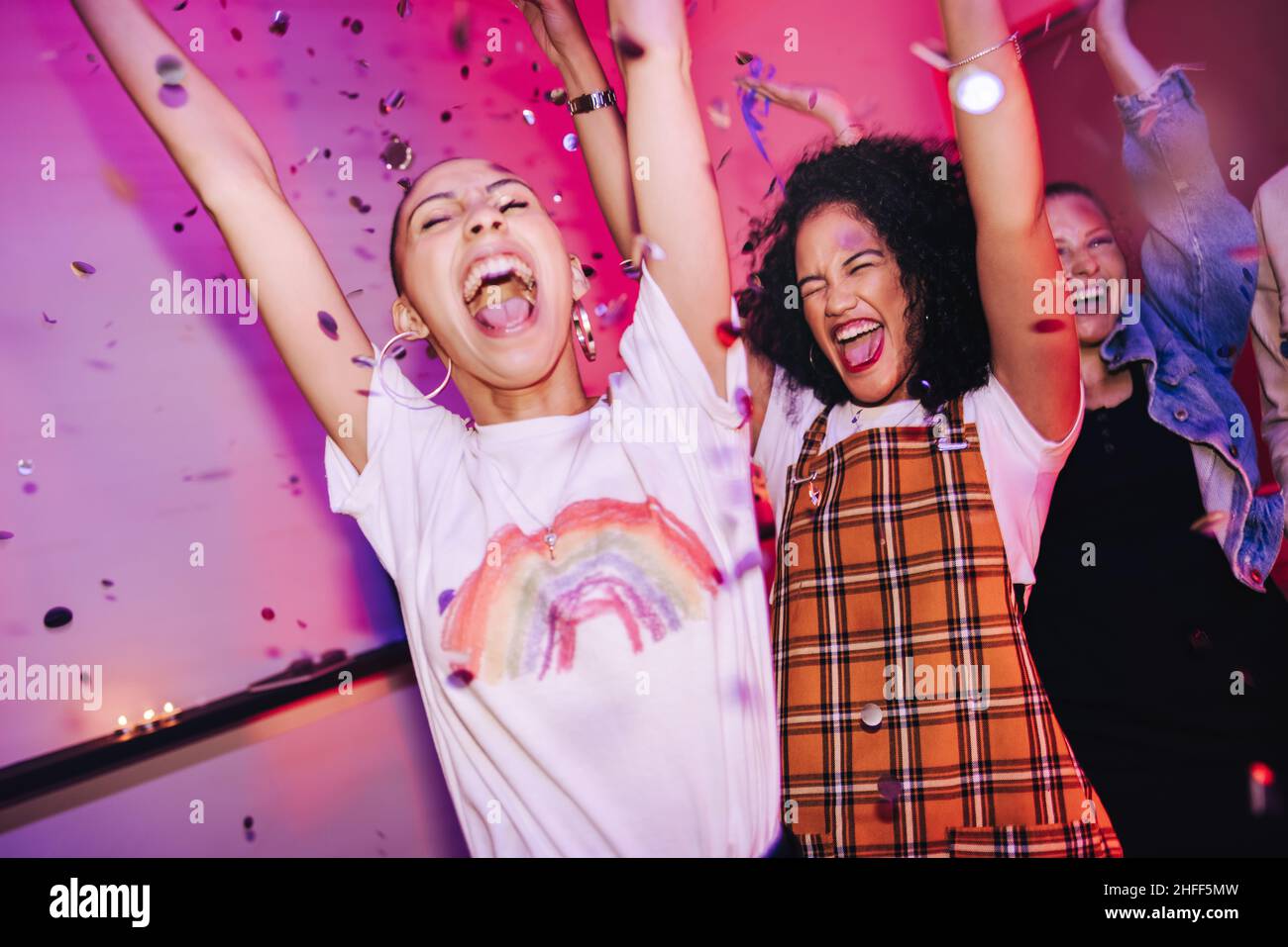Friends cheering happily at a house party. Cheerful female friends shouting for joy while standing under falling confetti. Group of vibrant young wome Stock Photo