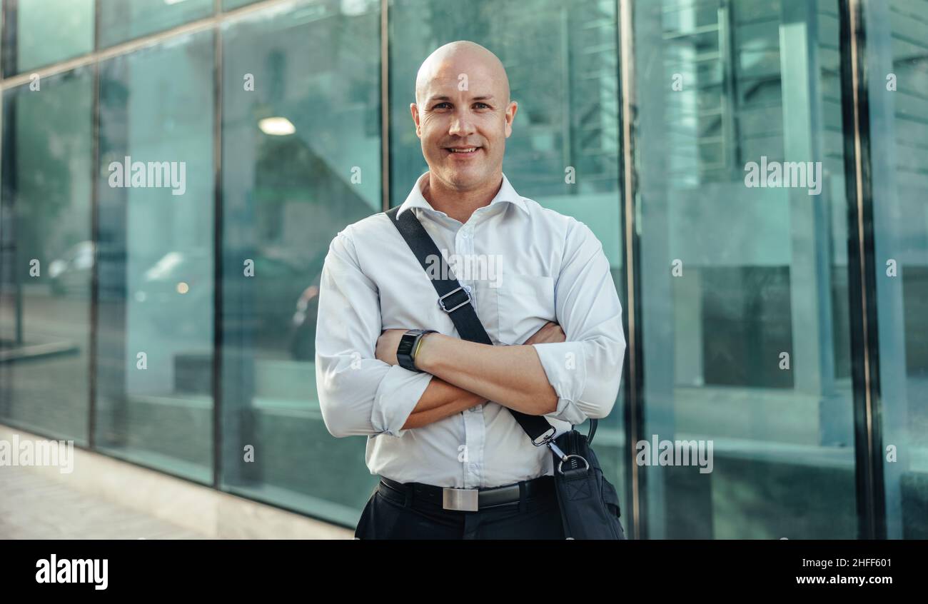 Successful businessman smiling at the camera cheerfully while standing in front of a high rise office building in the city. Mid-adult entrepreneur sta Stock Photo