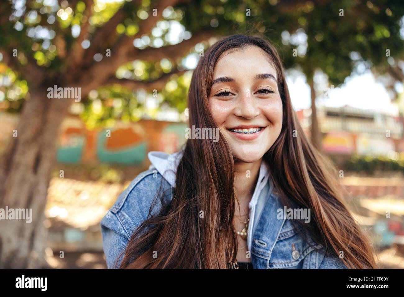 Smiling teenage girl looking at the camera outdoors. Happy young teenager wearing a denim jacket in the city. Female youngster sitting alone in an urb Stock Photo