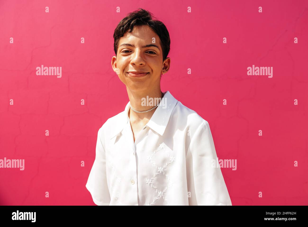 Smiling teenage boy standing against a pink background. Happy young queer boy looking at the camera while standing in a studio. Confident gay teenager Stock Photo