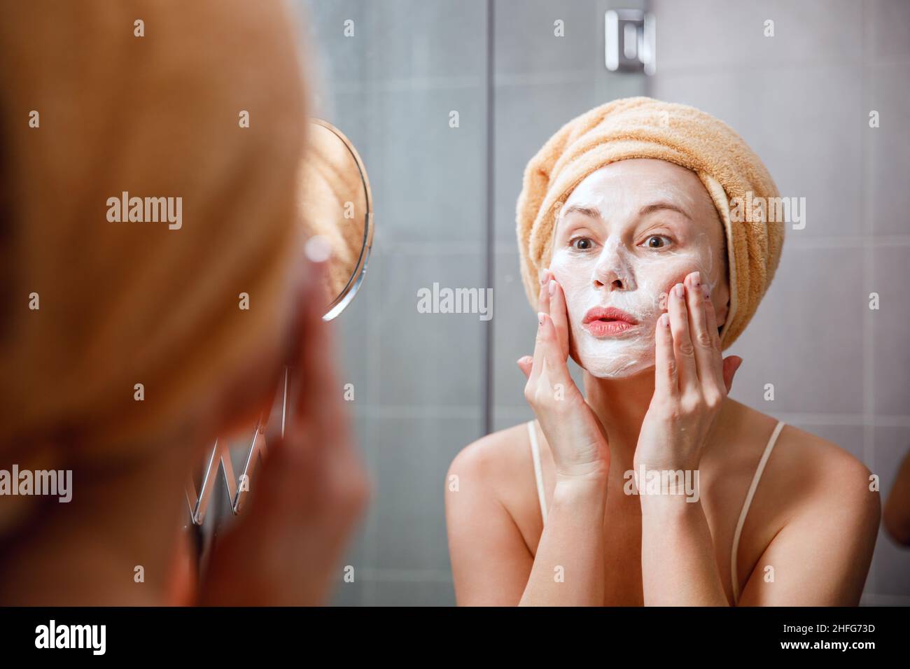A woman puts a cleansing mask on her face in the bathroom in front of the mirror. Skin care. Cosmetic spa treatments. Stock Photo
