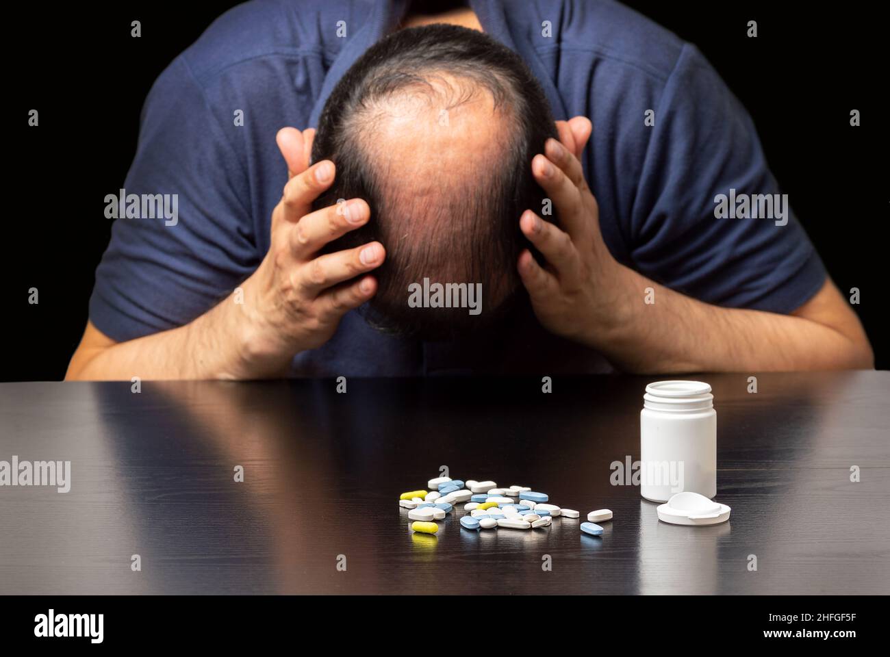 Close up of a depressed bald man next to a lot of pills on a table on a dark background Stock Photo