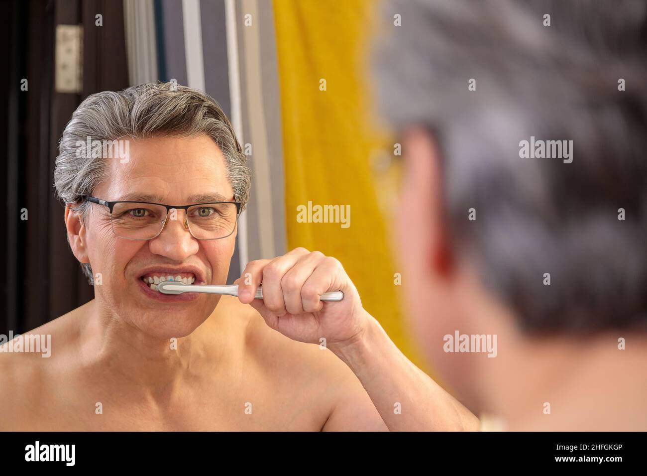 An adult gray-haired man brushes his teeth with a toothbrush and looks in the mirror. Stock Photo