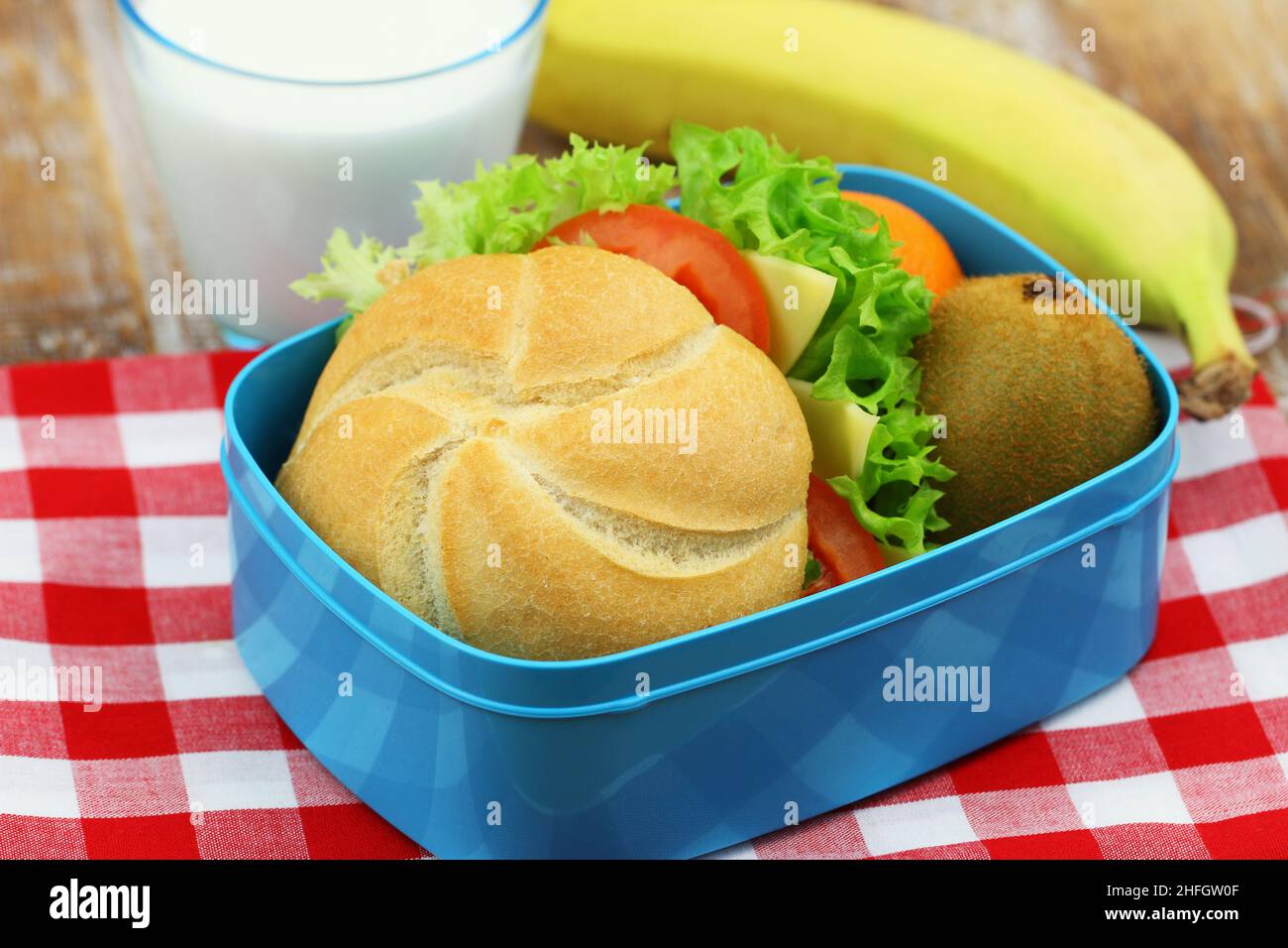 Healthy school lunchbox containing roll with cheese, lettuce and tomato, banana, kiwi fruit and glass of milk Stock Photo