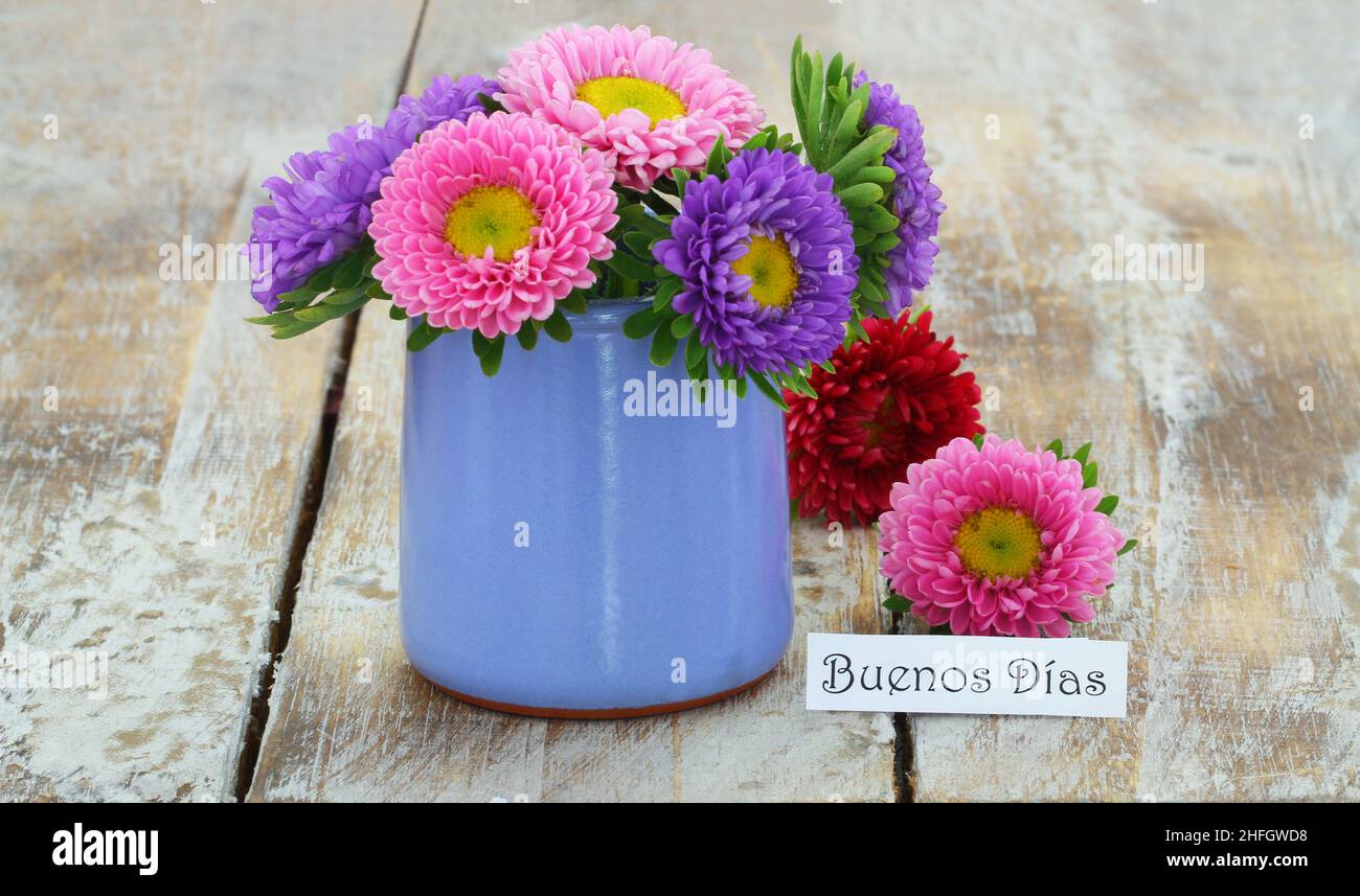 Buenos dias (good morning in Spanish) card with colorful daisies in blue vase on rustic wooden surface Stock Photo