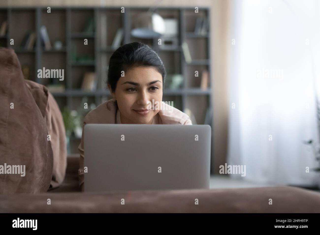 Pleasant happy young Indian woman using computer, resting on sofa. Stock Photo