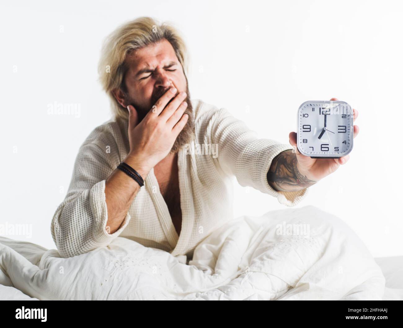 Sleepy bearded man with alarm clock. Yawning guy in bed with alarmclock. Early morning wake up. Awaking. Stock Photo