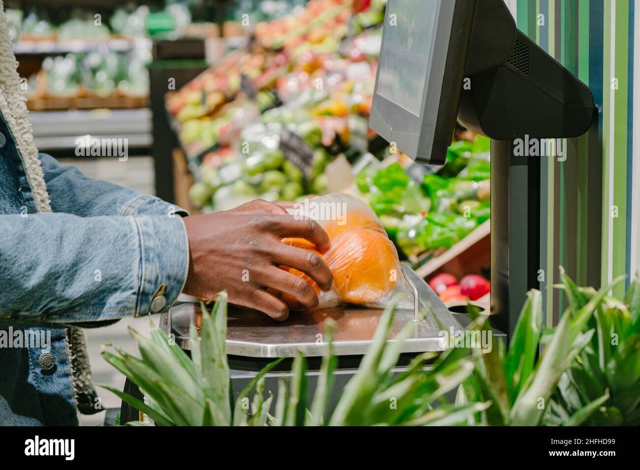 Stylish African-American guy in warm jacket with disposable mask weighs fresh oranges with digital scale in light supermarket Stock Photo