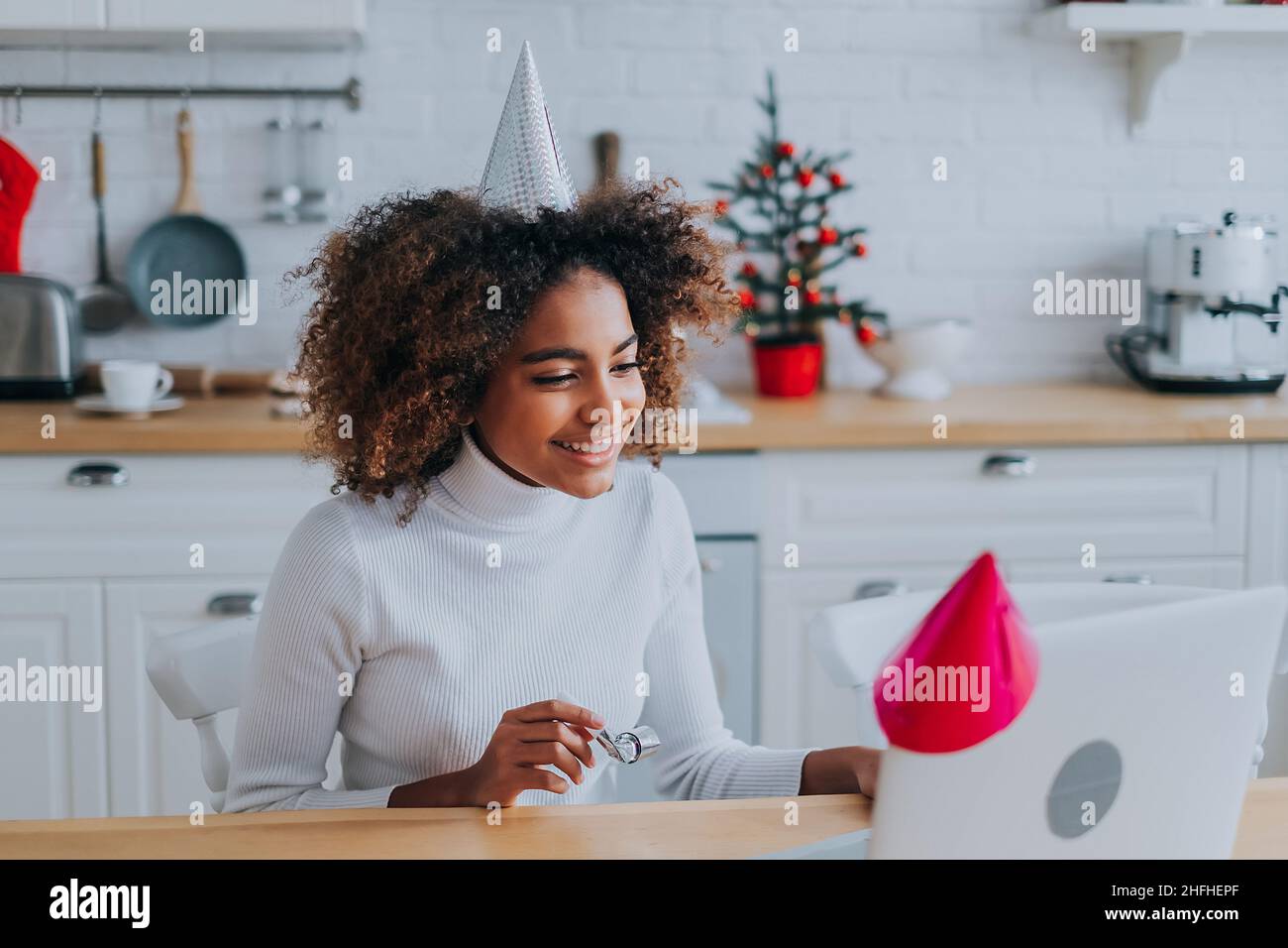 A beautiful black girl with a lush hairstyle in a festive hat on her head celebrates her birthday online by video chat sitting at home in the kitchen with a laptop, sends greetings online during quarantine. Stock Photo