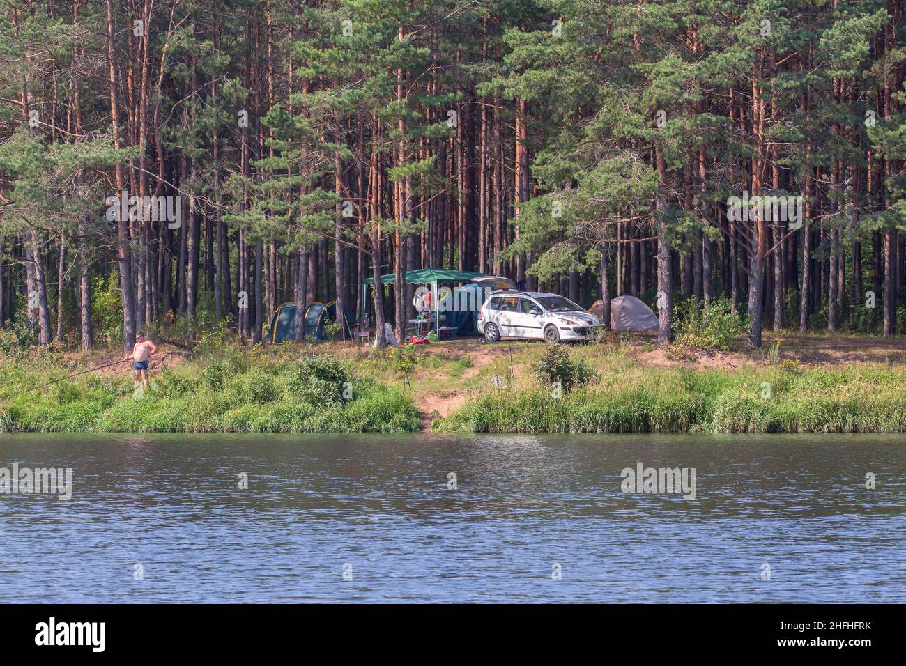 Tourists camped in the woods on the river bank: tents, awnings, and a car. A man catches fish. A sunny day in nature Stock Photo