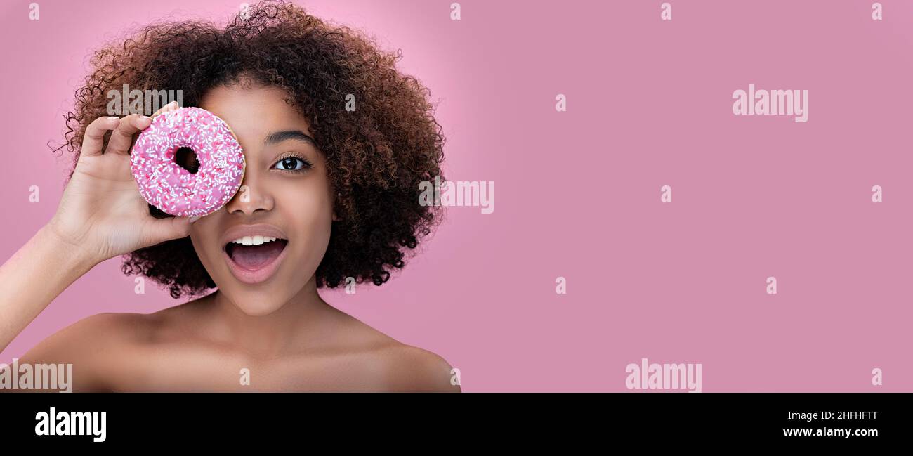 Portrait of a happy dark-skinned young woman covers one eye with a pink donut on a pink background, copy space Stock Photo