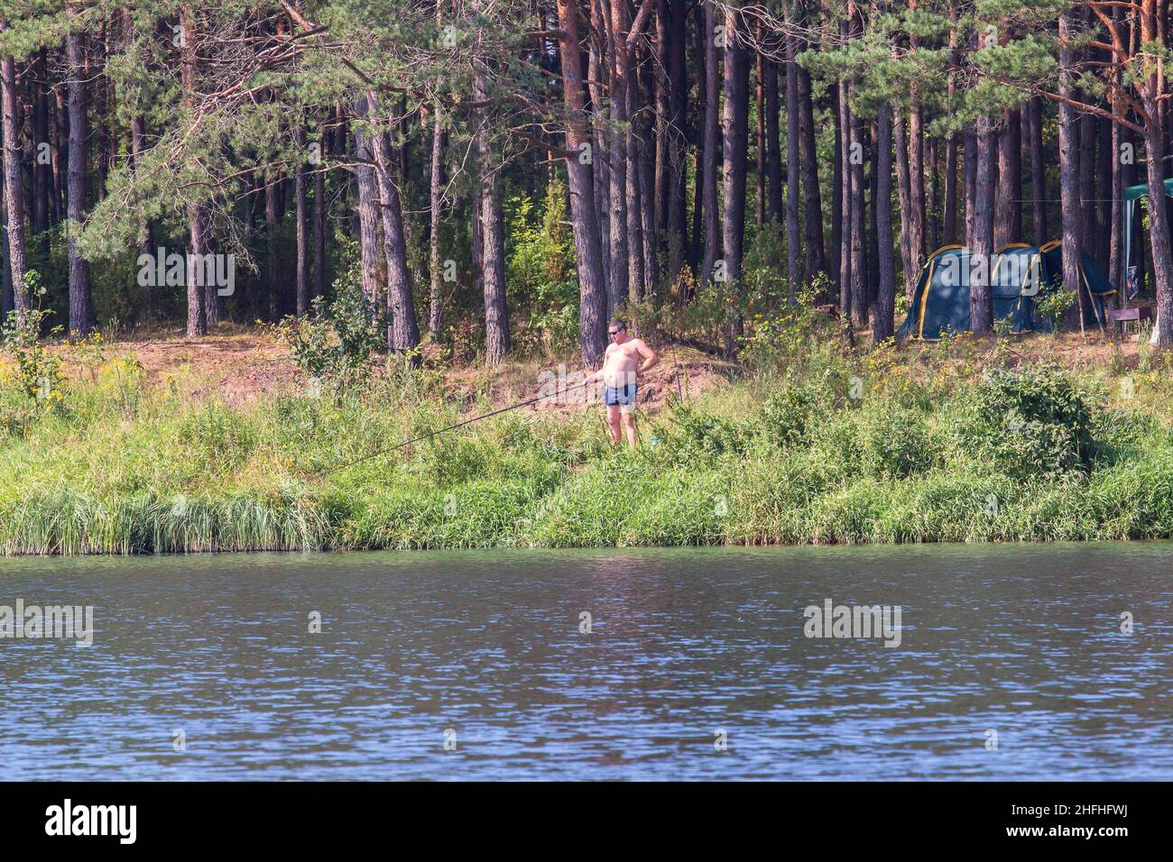Tourists camped in the woods on the river bank: tents, awnings. A man catches fish. A sunny day in nature Stock Photo