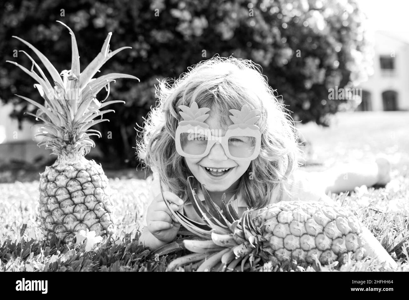 Boy with pineapple on head, plays with fresh tropical fruit outdoorst. Excited funny kid. Child face with amazed expression. Kids summer party. Stock Photo