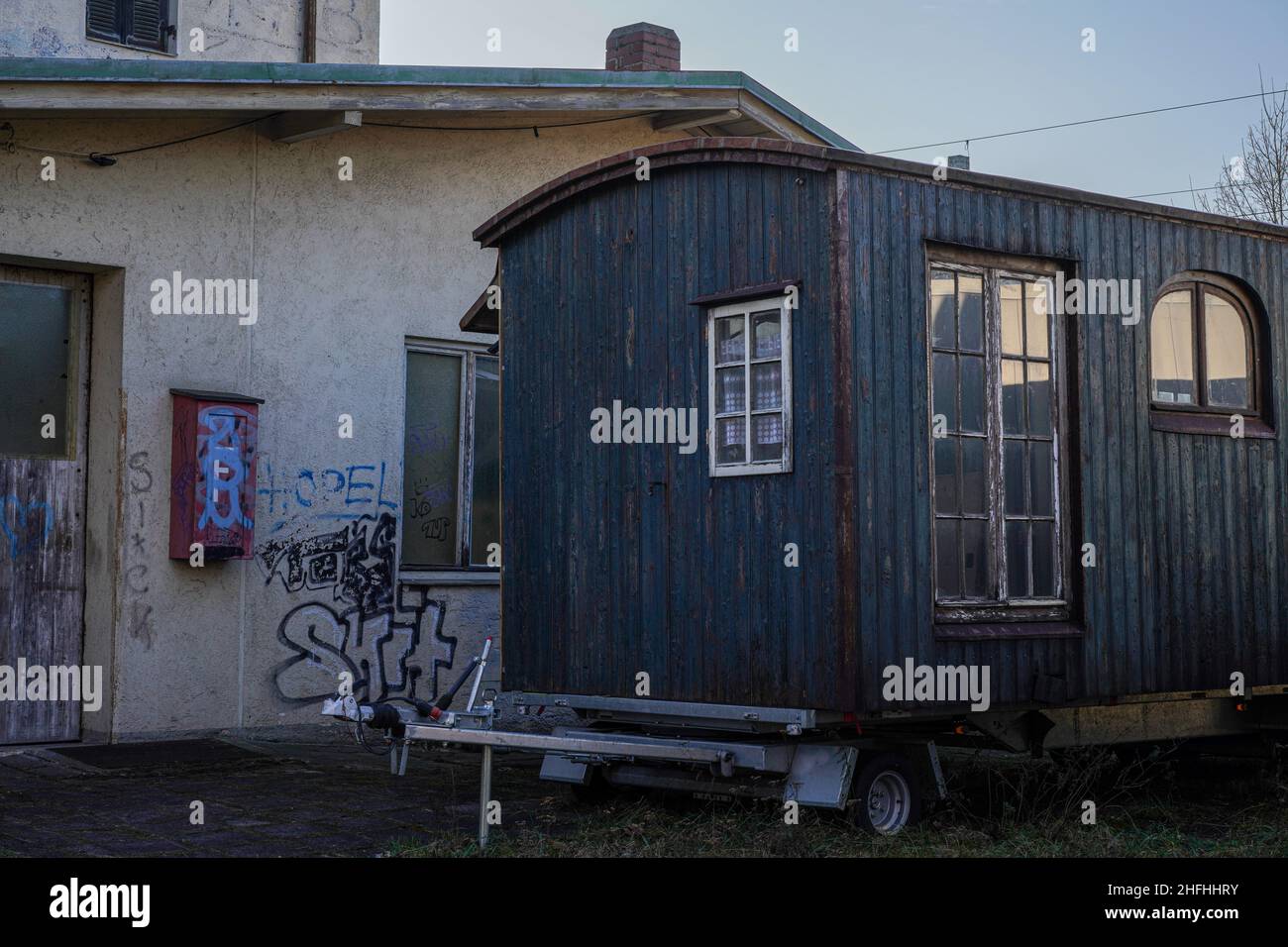 An old wooden caravan trailer with three windows, one of them large, is parked in front of an uninhabited house. Stock Photo